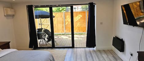 Un dormitorio con una cama, un televisor montado en la pared y un ventanal corredero que da a un patio exterior con mesa y sillas.