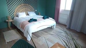 A bedroom with a bed, a round wooden headboard, a woven wall panel, a window with curtains, a green chair, and a wooden floor. - Gaby's Little House (Saint-Hilaire-de-Riez)