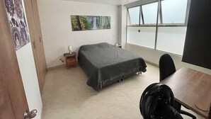Un dormitorio con cama, escritorio, silla, ventana con vista, un cuadro en la pared y un ventilador.