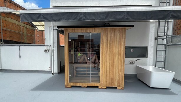Una sauna moderna al aire libre con revestimiento de madera, puerta de vidrio y una bañera blanca.