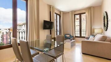 A modern hotel room with a glass dining table, a sofa, a TV, and a balcony with a view.