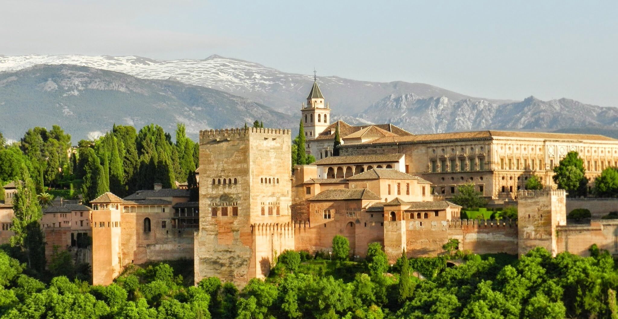 Un edificio histórico con una torre destacada, rodeado de exuberante vegetación y montañas al fondo.