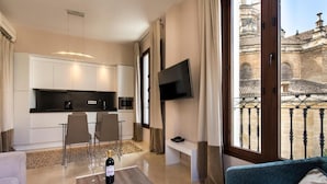 A modern living room with a sofa, a coffee table with a bottle, a TV mounted on the wall, and a balcony with a view of historic architecture. - Siloe Plaza Apartments (Granada)