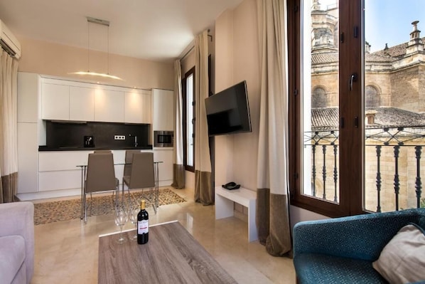 A modern living room with a sofa, a coffee table with a bottle, a TV mounted on the wall, and a balcony with a view of historic architecture.