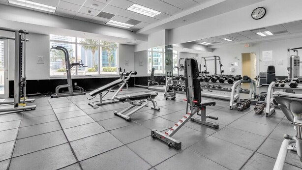Un gimnasio con diversos equipos de ejercicio, amplias ventanas y un reloj en la pared.