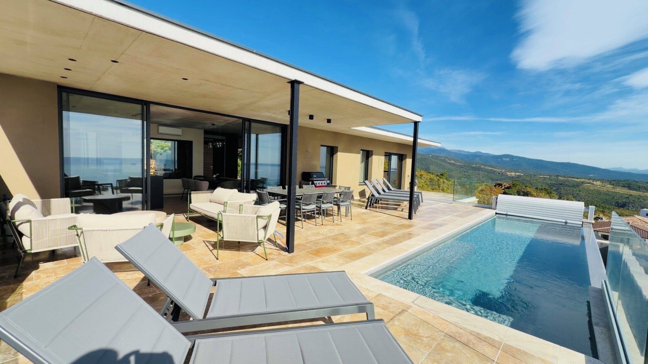 A modern outdoor pool area with lounge chairs, a barbecue, and a view of the mountains.