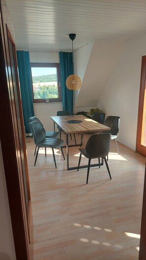 Ein Essbereich mit einem Holztisch, vier Stühlen, einer Pendelleuchte und Blick durch ein fensterbepflanztes Fenster.