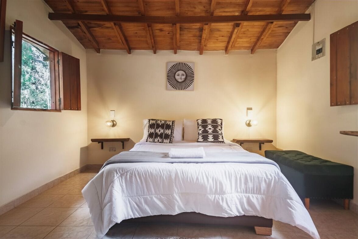 Un dormitorio con techo de madera, una cama con ropa de cama blanca, dos mesitas de noche con lámparas de pared y un banco verde.