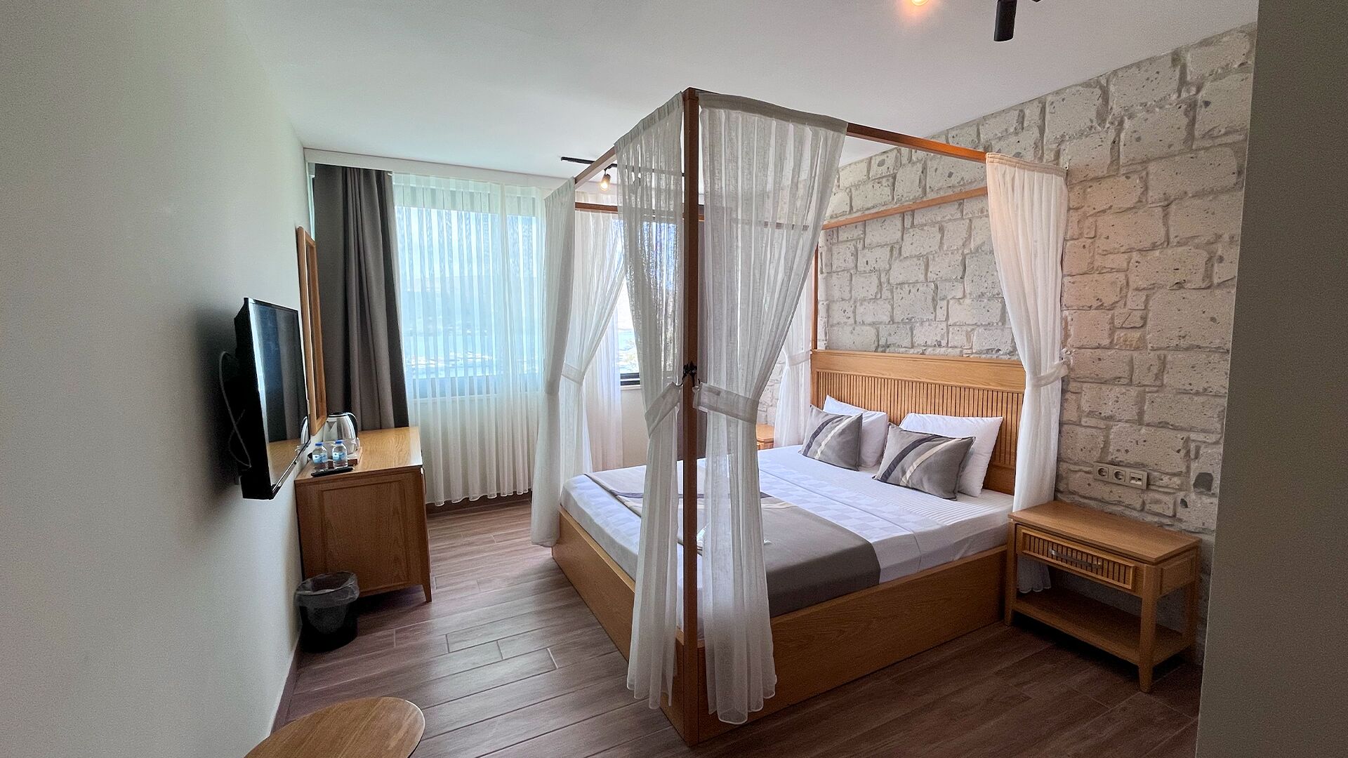 A bedroom with a four-poster bed, a wooden nightstand, a bedside table, a TV mounted on the wall, and a brick-patterned wall.