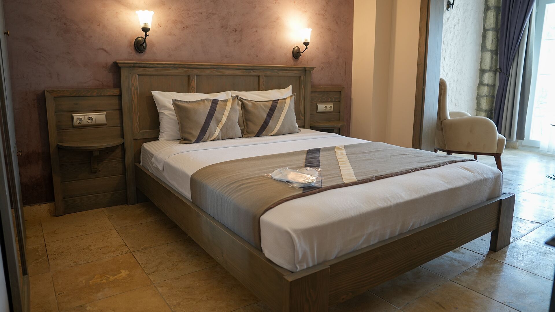 Une chambre d’hôtel avec un lit en bois, deux tables de chevet, une chaise et un luminaire fixé au mur.