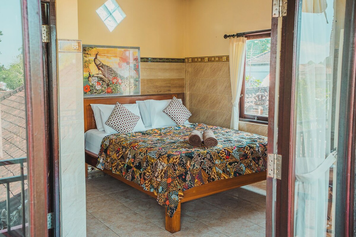 Une chambre à coucher comprenant un lit, un ventilateur de plafond, une fenêtre avec des rideaux et un mur orné d’un élément décoratif.