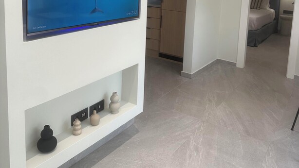 Una camera moderna con televisione a schermo piatto montata a parete, un armadio in legno, un letto con comodino e un piccolo ripiano con oggetti decorativi.