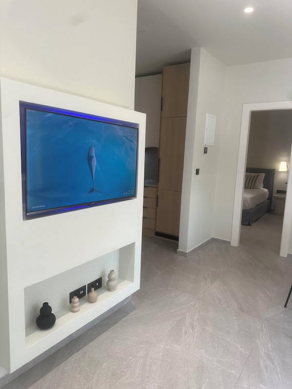 Ein modernes Zimmer mit einem wandmontierten Flachbildfernseher, einem Holzschrank, einem Bett mit Nachttisch und einer kleinen Ablage mit Dekoration.