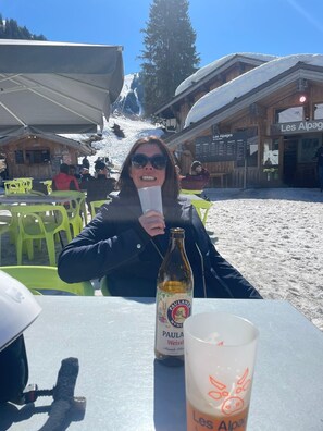 En person sitter vid ett bord med en ölflaska och ett glas. I bakgrunden syns ett snötäckt berg och en träbyggnad med skylten ”Les Alpage”.