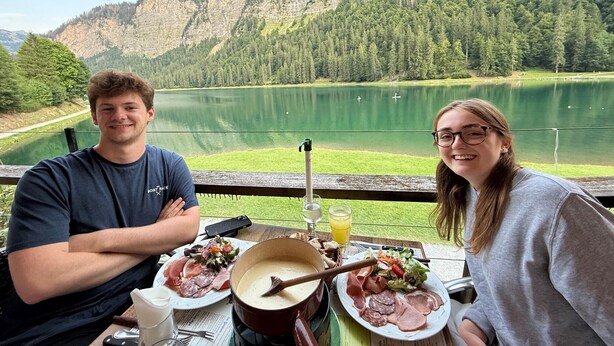 Zwei Personen sitzen an einem Tisch mit einer Mahlzeit und blicken auf einen See und Berge.