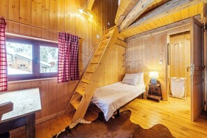 Ein Zimmer einer Holzhütte mit einem Bett, einem fenster mit Vorhängen, einer Leiter, einem kleinen Tisch und einer Tür.