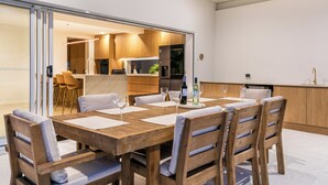 A dining area with a wooden table and chairs, a kitchen in the background, and a sliding glass door leading to an outdoor space. - Bliss On The Avenue - Refined living (Burleigh Waters)
