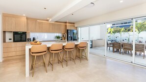 A modern kitchen with a central island, bar stools, and a dining area with a view. - Bliss On The Avenue - Refined living (Burleigh Waters)