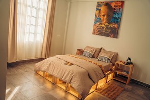 Quarto com cama, cabeceira de madeira, mesa de cabeceira, pôster na parede e janela com cortinas.