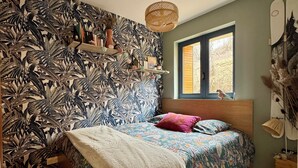 A bedroom with a bed, a window, a wall with a tropical leaf pattern, a shelf with decorative items, and a hanging light fixture. - "SIGI" Cozy apartment at the foot of the Ceüse cliff (Sigoyer)