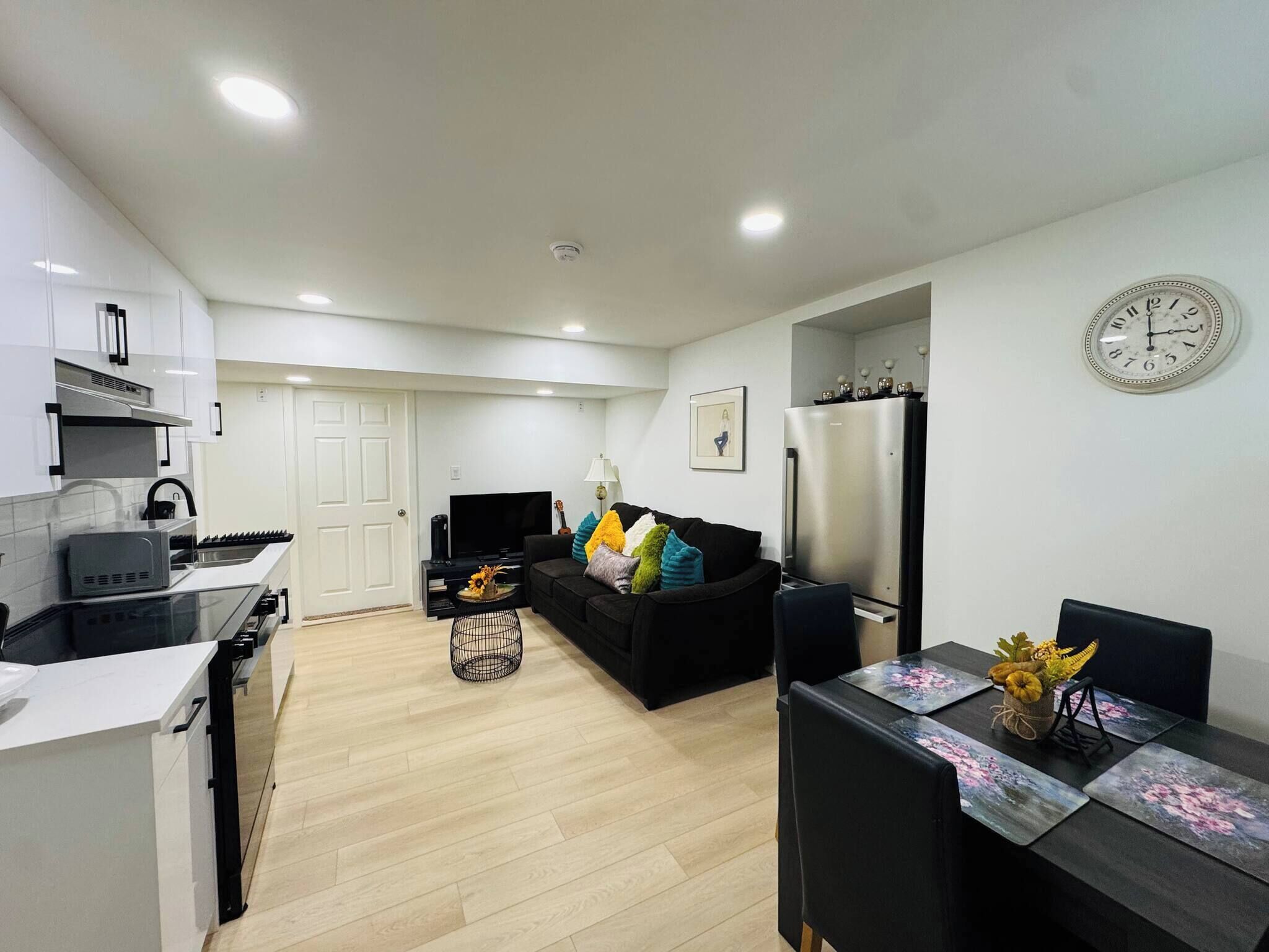 A modern kitchen with white cabinets, a black countertop, and a built-in oven. There is a black sofa with colorful cushions, a dining table with floral placemats, and a wall clock.