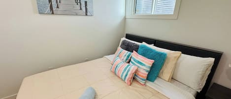 A bedroom with a bed, pillows, a rolled towel, a painting of a pier, and a window.