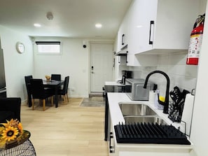 Una cuina compacta amb microones, rentamicroplates i aixeta negra. Hi ha una zona de menjador amb taula i cadires, i una porta blanca al fons.