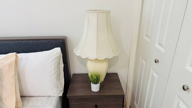 Un letto con biancheria bianca, un comodino in legno scuro con una lampada e una piccola pianta, e una porta bianca.