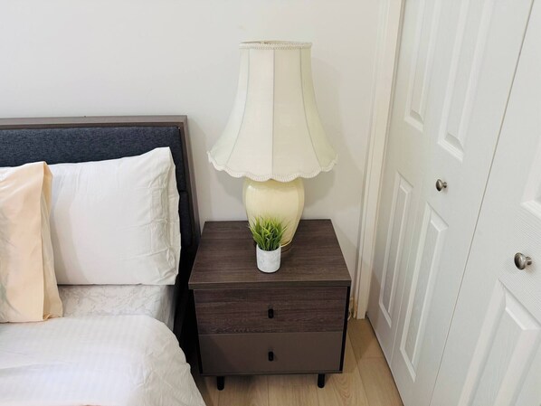 A bed with white bedding, a dark wooden nightstand with a lamp and a small plant, and a white door.