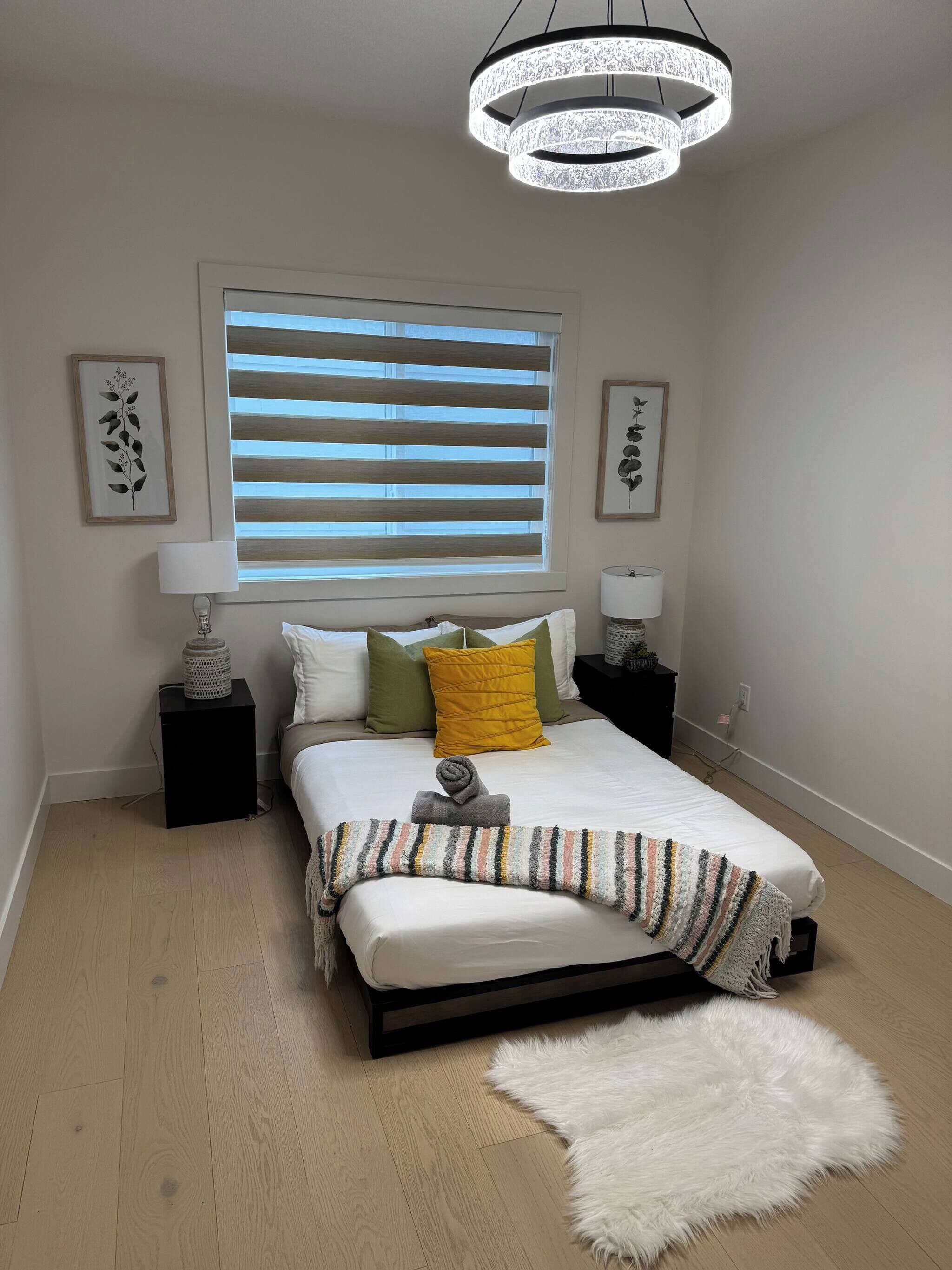 A bedroom with a bed, a circular ceiling light, a window with blinds, bedside tables, and wall art.