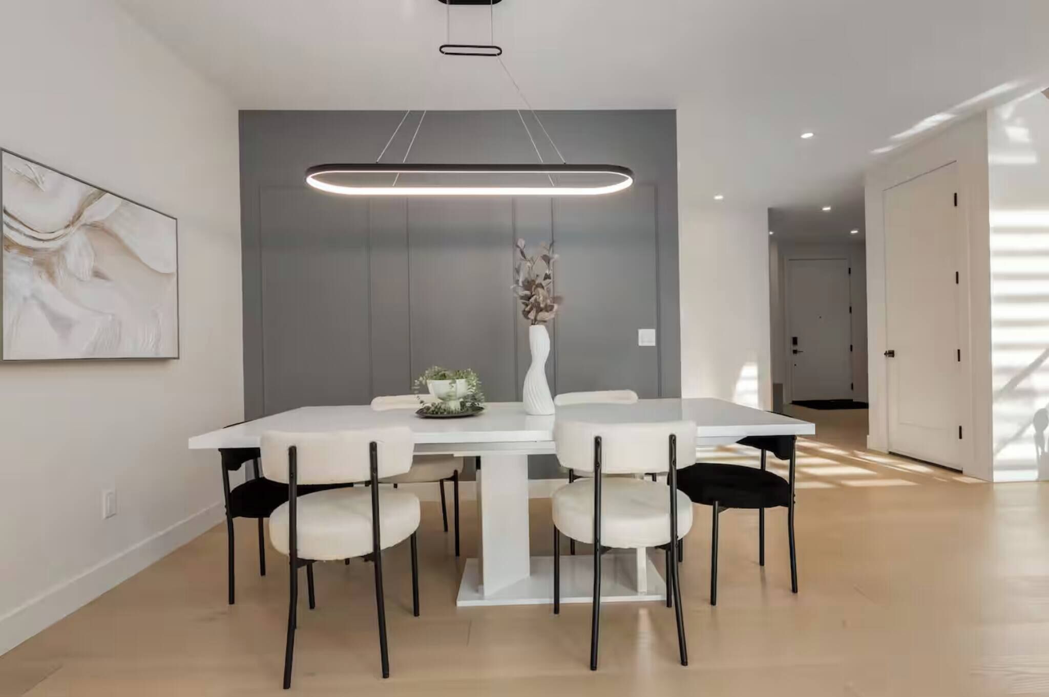 Un espace repas moderne avec une table blanche, des chaises noires, un luminaire suspendu et un vase rempli de fleurs.