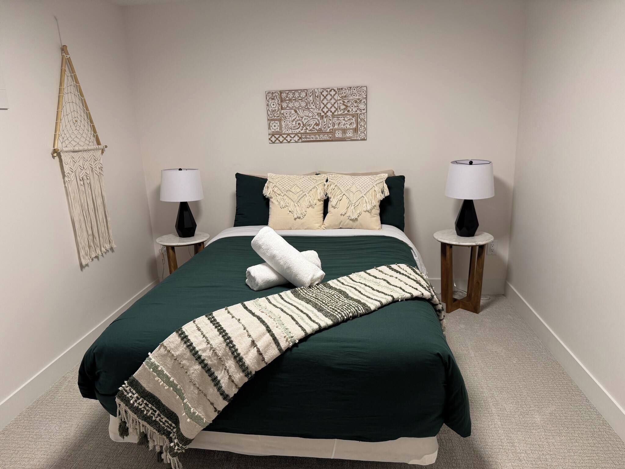 Un dormitorio con una cama, dos mesitas de noche, un colgador de macramé, un adorno de pared y una manta con estampado.