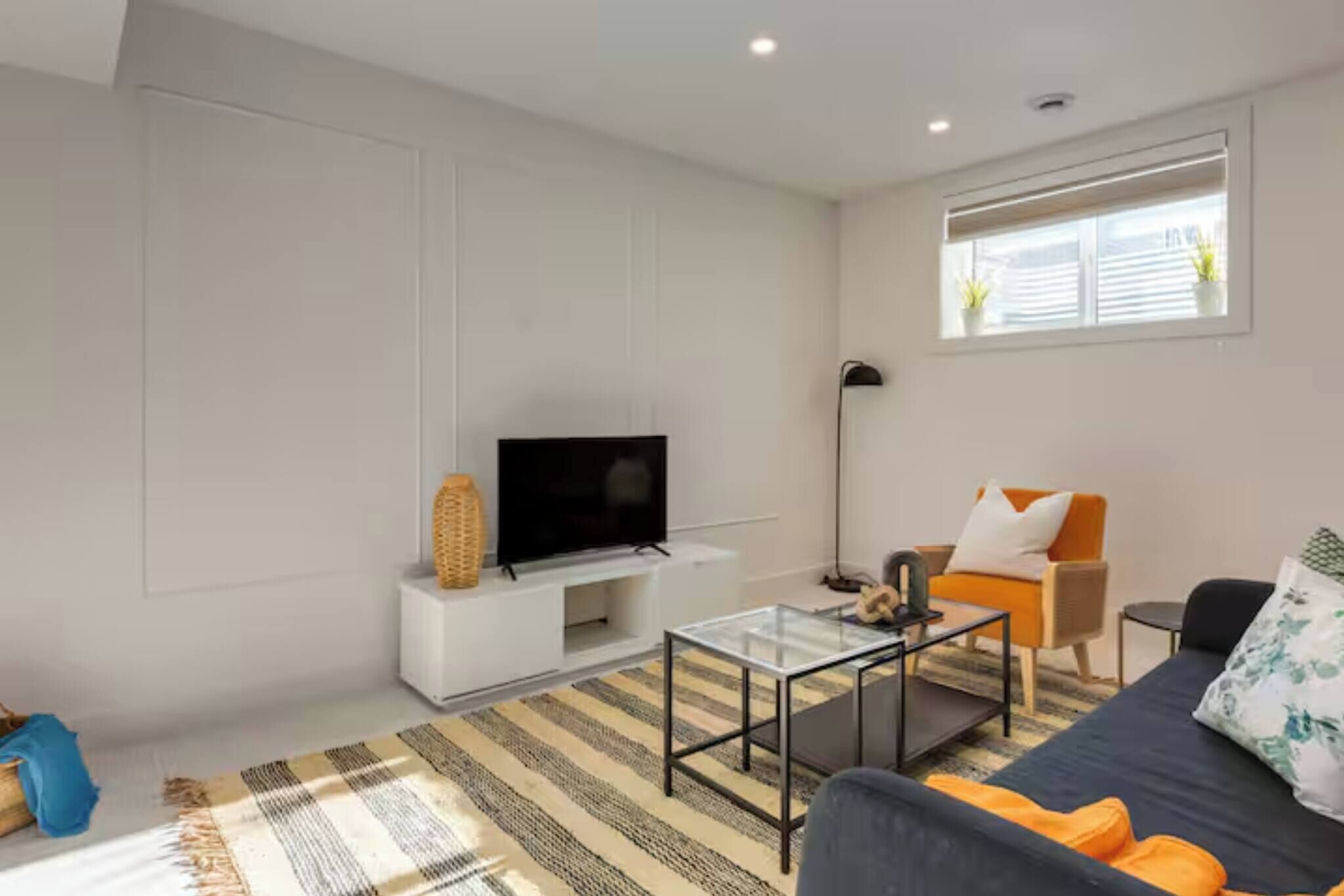 Una sala de estar moderna con una televisión de pantalla plana, una mesa de centro de vidrio, un sofá y una ventana con persianas.