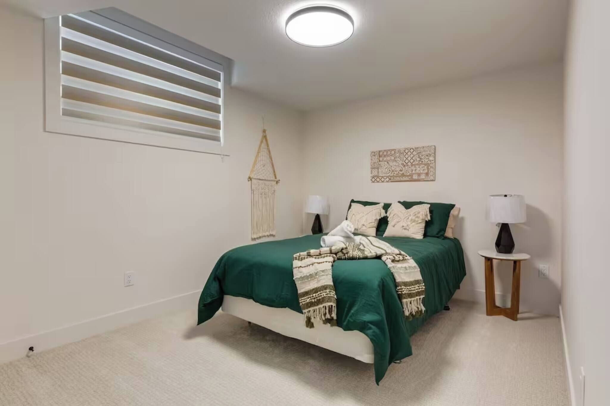 A bedroom with a bed, bedside lamps, a wall-mounted light fixture, a window with blinds, and a decorative wall hanging.