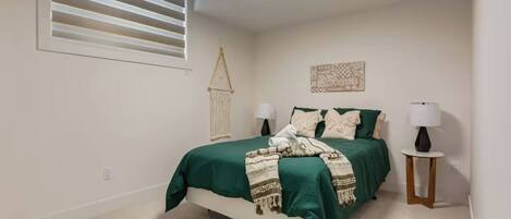 A bedroom with a bed, bedside lamps, a wall-mounted light fixture, a window with blinds, and a decorative wall hanging.
