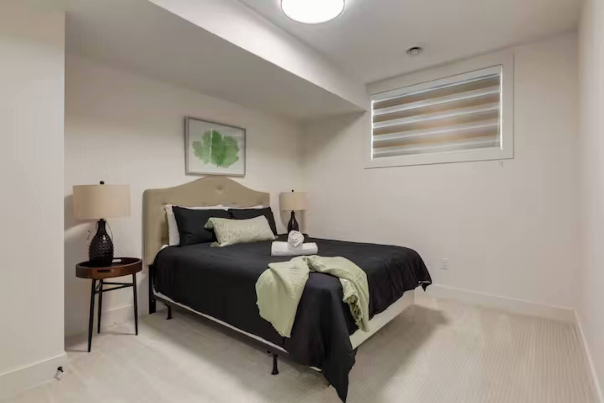 A bedroom with a bed, two bedside tables, a wall-mounted light fixture, a window with blinds, and a framed artwork on the wall.