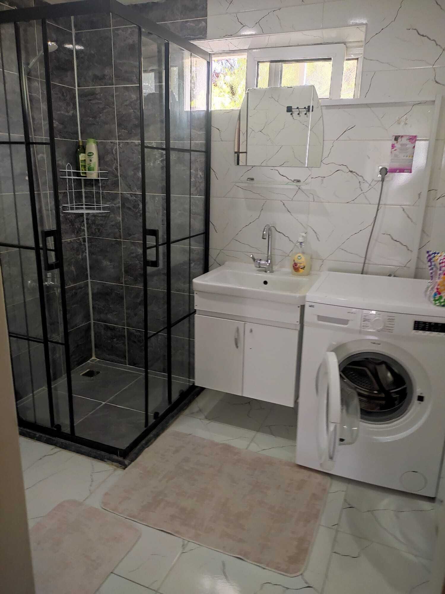 Un baño con ducha envuelta en vidrio, un lavamanos con espejo y una lavadora.
