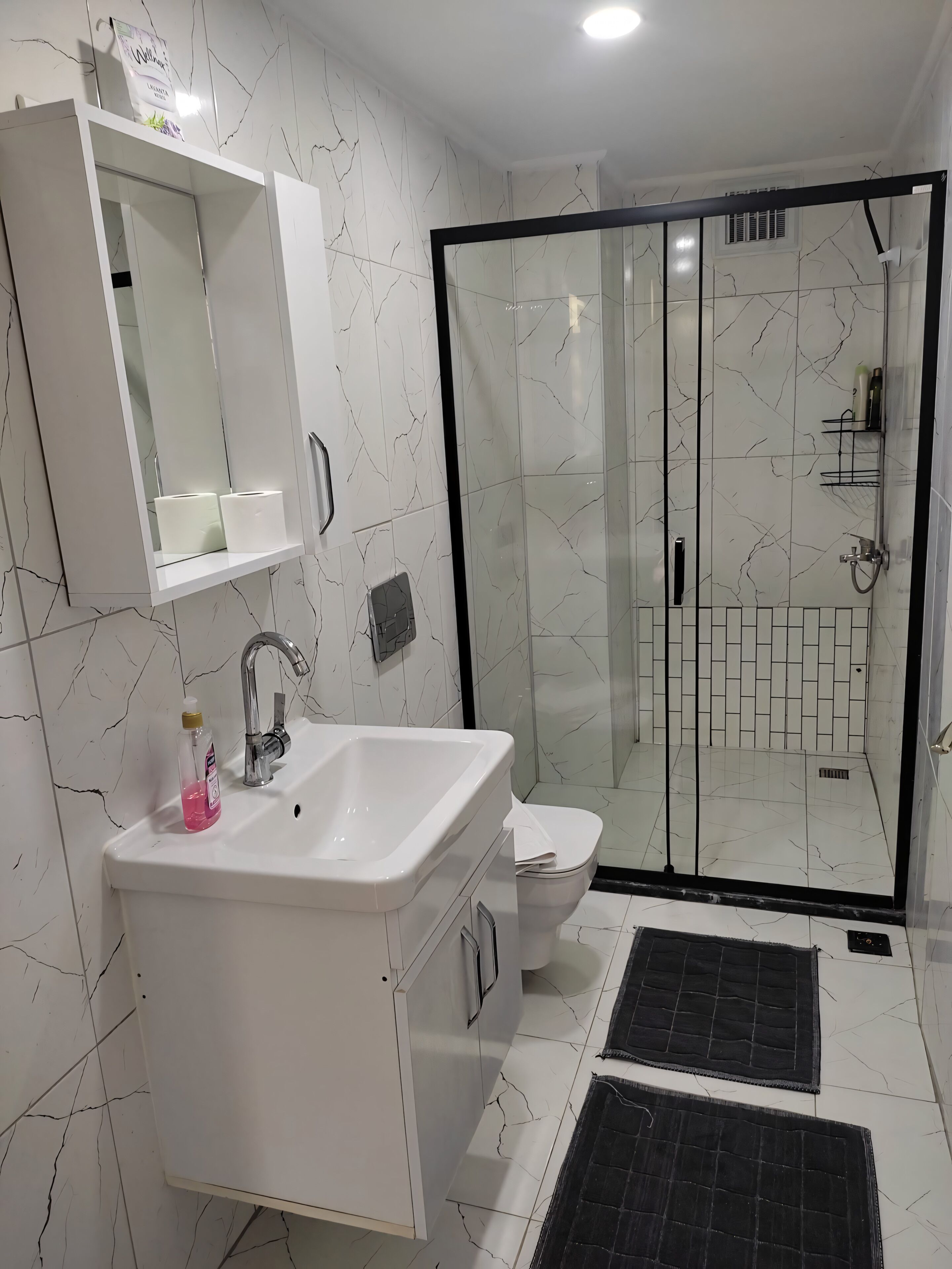 Un baño moderno con un tocador blanco, cabina de ducha de vidrio y un tapete negro en el suelo.