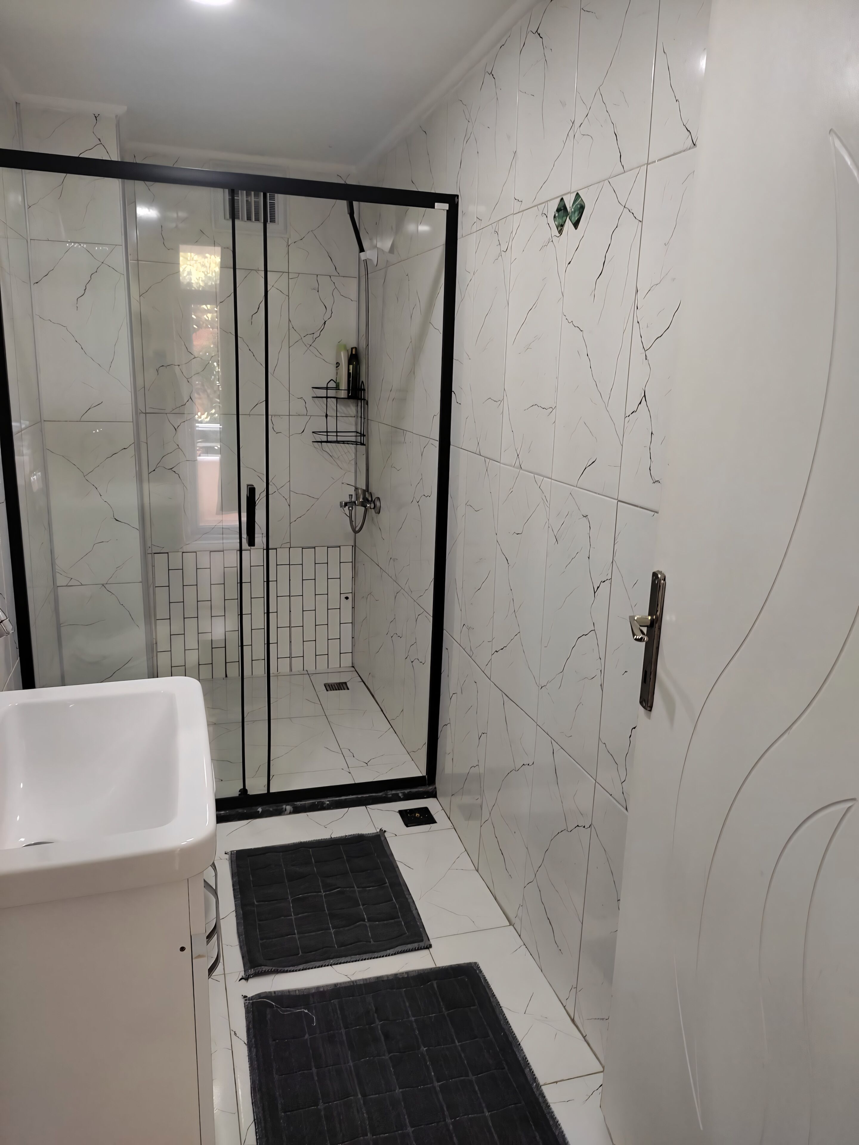 A bathroom with white marble tiles, a glass shower enclosure, a white bathtub, and a black mat on the floor.