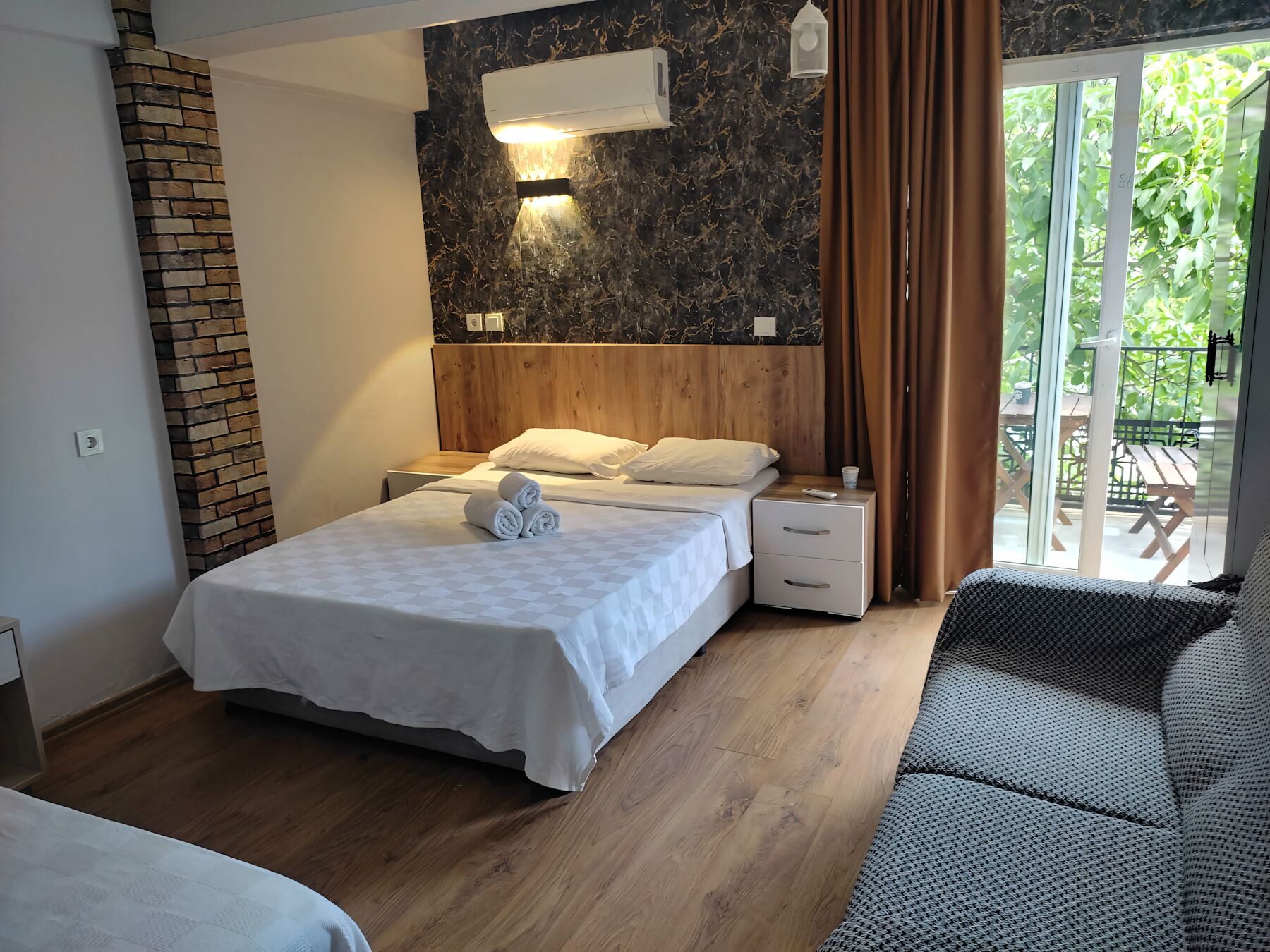 A hotel room with a bed, wooden headboard, nightstands, and a view of greenery through a glass door.