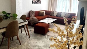 Una sala de estar con sofá, reposapiés, mesa de centro, mesa de comedor con sillas y una planta en maceta.