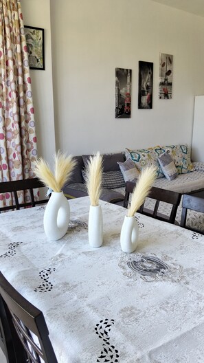 Una mesa de comedor con mantel de encaje, tres jarrones blancos con plumas de pampa, un sofá con cojines estampados y obras de arte en la pared que representan paisajes urbanos.