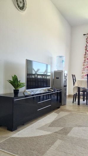A living room with a flat-screen TV, a black entertainment unit, a potted plant, a water dispenser, and a dining area with a table and chairs.