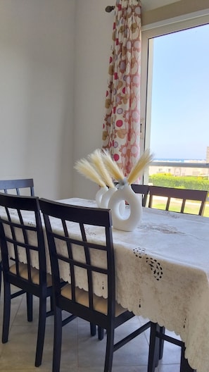 A dining table with chairs, a floral curtain, and a view of the sea.