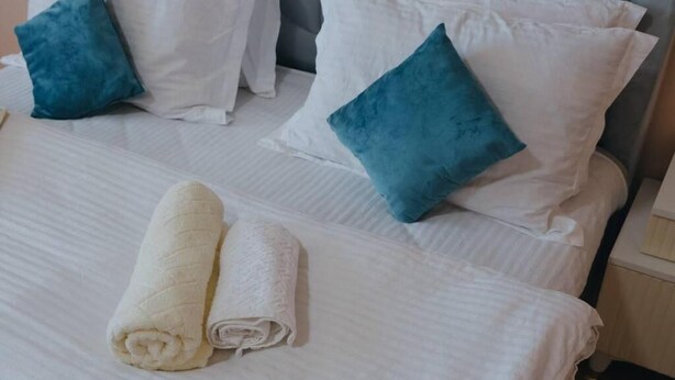 Una cama con ropa de cama blanca, dos almohadas azules y dos toallas enrolladas.