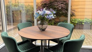 Une table ronde entourée de quatre chaises vertes, un vase rempli de fleurs et une vue sur un jardin à travers une paroi de verre.