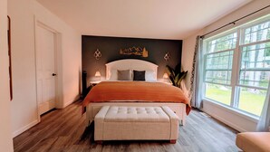 Ein Schlafzimmer mit einem großen Bett, einer Bank, einem Nachttisch, einer Wandleuchte und einem Fenster mit Blick auf Bäume.