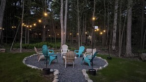 Ein runder Sitzbereich mit Adirondack-Stühlen und einem Tisch, umgeben von einem Kiesweg und Bäumen, mit Lichterketten darüber.