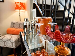 A bar area with various glassware and bottles, a sofa with patterned cushions, and a staircase in the background. - 2BR Condo | Pool & Hot Tub | Downtown Palm Springs | Mountain Views (Palm Springs)