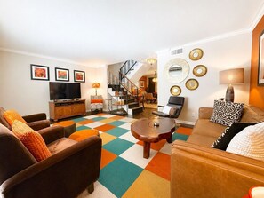 Ein Wohnzimmer mit Sofa, Sesseln, einem Couchtisch, einem Fernseher und einer Treppe, die in einen Essbereich führt.